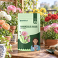 A stand-up pouch of Paankhuri Ranunculus Bulbs (mix colour) is centered on a wooden table surrounded by potted flowers, including pink, red, and yellow ranunculus, with a small gardening trowel in the foreground.