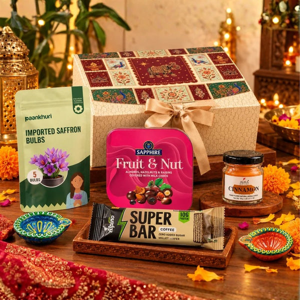 A festive gift basket arrangement on a wooden table, featuring a large patterned gift box tied with a beige ribbon, a package of Paankhuri imported saffron bulbs, a pink Sapphire Fruit & Nut tin, an Alvaro Super Bar coffee snack, and a small cinnamon jar, surrounded by decorative lamps and flowers.