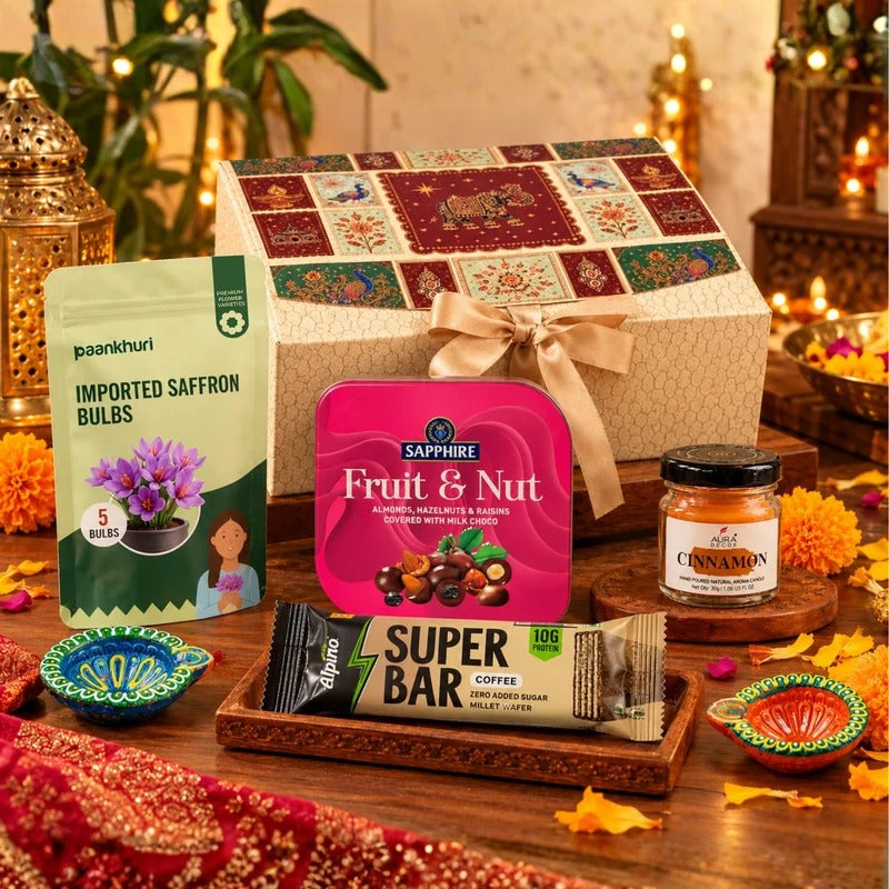 A festive gift basket arrangement on a wooden table, featuring a large patterned gift box tied with a beige ribbon, a package of Paankhuri imported saffron bulbs, a pink Sapphire Fruit & Nut tin, an Alvaro Super Bar coffee snack, and a small cinnamon jar, surrounded by decorative lamps and flowers.